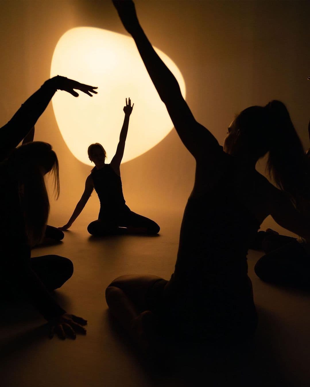 Silhouettes of people seated on the floor with arms raised, lit by a large abstract light.