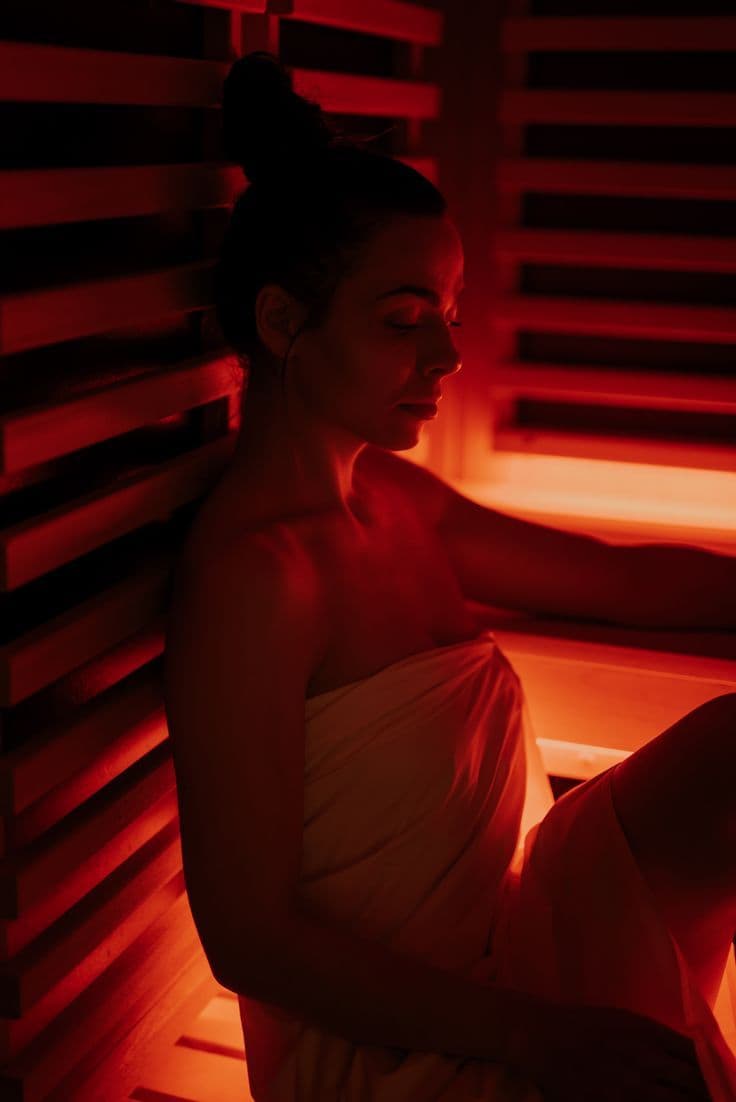 Woman wrapped in a towel relaxing inside a red-lit infrared sauna room.