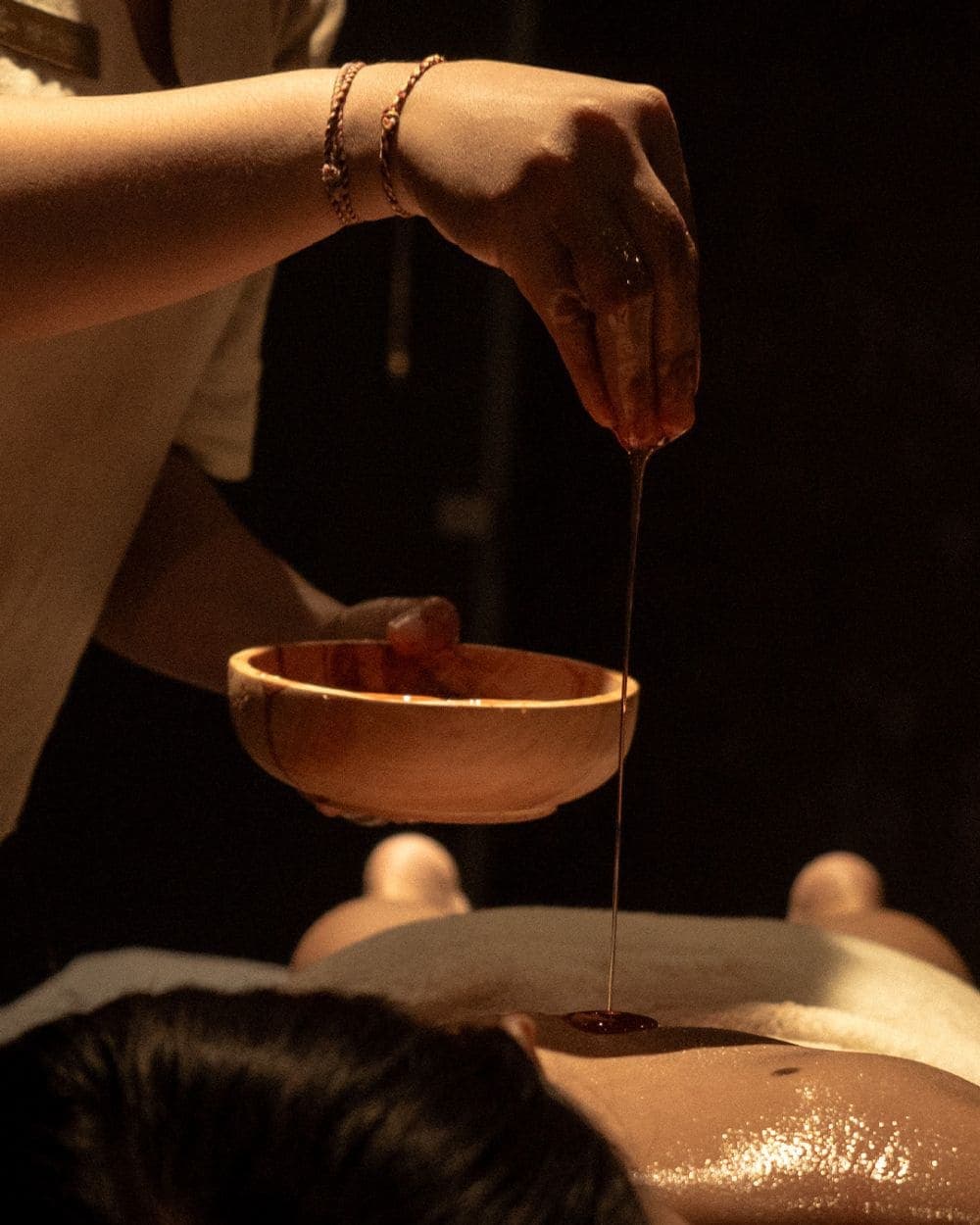 A hand pouring warm oil from a wooden bowl onto a person's back during a massage.