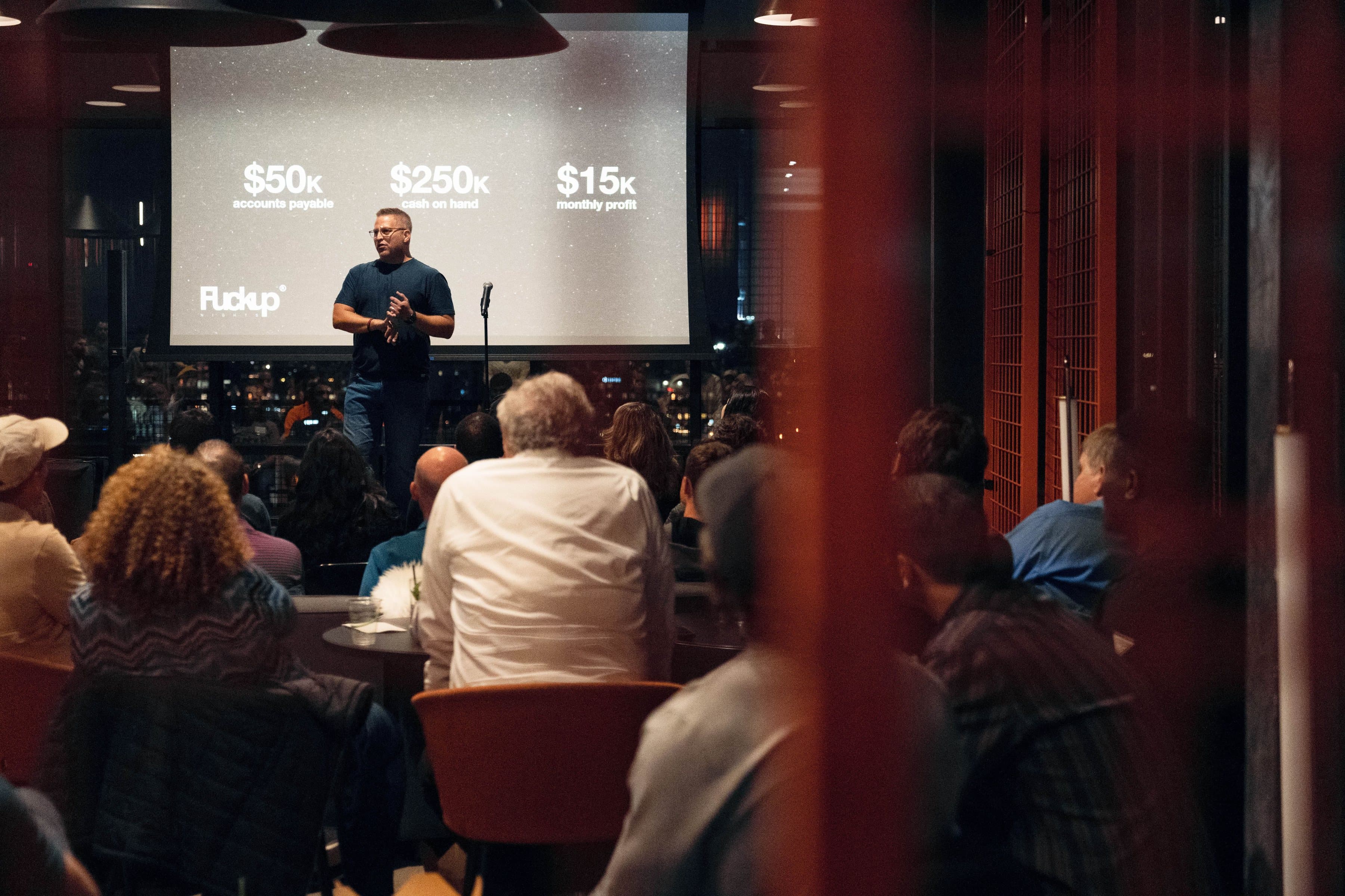 A speaker presenting to a seated audience in front of a large lit screen.